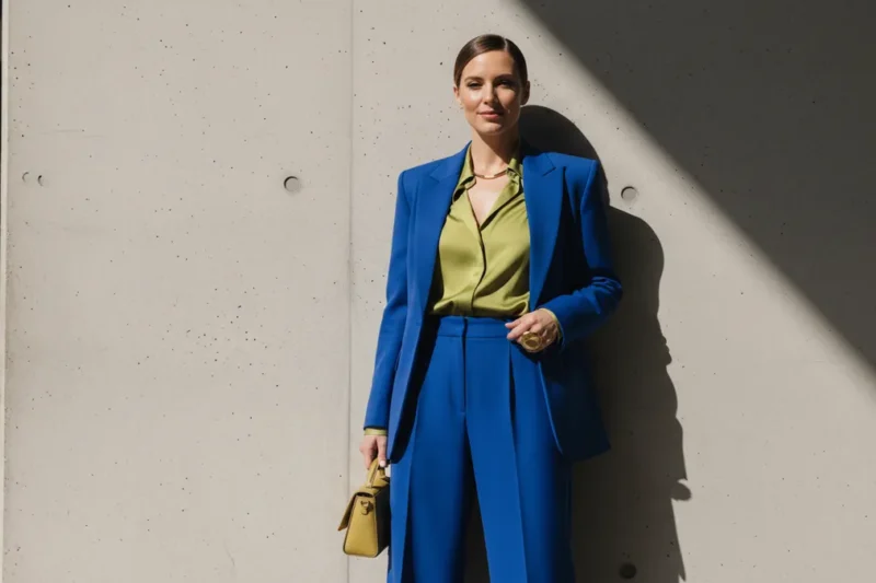 Woman in a bright blue pantsuit and lime blouse stands against a concrete wall in sunlight, holding a yellow handbag.